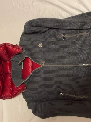 Moncler dunväst med huva, röd/grå - Säljer en snygg dunväst från Moncler i storlek M. Västen har en röd quiltad framsida och huva, samt grå stickade sidor och rygg. Utrustad med dragkedja och två fickor med zip. Moncler-logga på bröstet. Perfekt för dig som vill ha något både varmt och stilrent.