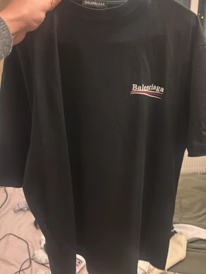 Svart Balenciaga t-shirt med tryck - Säljer en svart t-shirt från Balenciaga med stort vitt och rött logotryck på bröstet. Klassisk rund hals och korta ärmar. Materialet känns som mjuk bomull och passformen är loose, perfekt för en chill och trendig look.