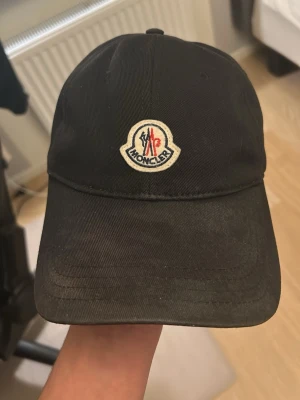 Svart Moncler keps med brodyr - Säljer en svart keps från Moncler med klassisk böjd skärm och broderad logga framtill. Justerbar rem bak med metallspänne och röd-vit-blå detalj. Snygg och enkel design som passar till det mesta. Den är dock solblekt, vet inte om man kan ta bort de på något sätt hör av er gärna om ni har funderingar 👍