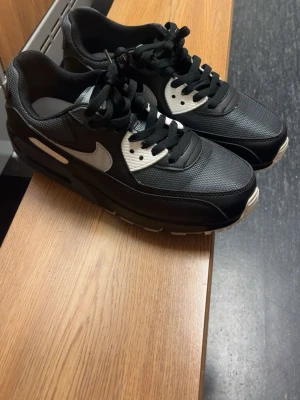 Nike Air Max 90 svart/vit EUR 43 - Nike Air Max 90 sneakers i svart och vit med grå detaljer. Skorna har en klassisk siluett med snörning, platt sula och rund tå. Ovandelen är i syntet och mesh, med tydlig Air Max-dämpning i sulan. Snygg kontrast mellan svart och vitt, perfekt för dig som gillar stilrena sneakers.