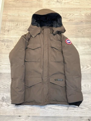 Canada Goose jacka - Brun dunjacka från Canada Goose med huva, fyra stora fickor framtill och klassisk logga på ärmen. Jackan har en rak passform och är fodrad med svart innerfoder. Perfekt för kalla vinterdagar och har snygga detaljer som tryckknappar och dragkedja.