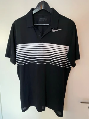 Golftröja från Nike Golf standard fit - Svart golftröja från Nike Golf med vita horisontella ränder över bröstet och ryggen. Toppen har korta ärmar, klassisk krage och två knappar framtill. Tillverkad i Dri-Fit polyester som andas och håller dig torr under golfrundan.