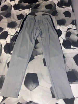 Grå träningsbyxor från SOC - Säljer ett par grå träningsbyxor från SOC med svarta detaljer längs sidorna. Byxorna har elastisk midja med snörning och är gjorda i ett syntetmaterial som är perfekt för sport och träning. Passformen är normal och byxorna har raka ben.