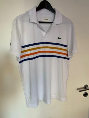 Lacoste golf-piké stl M - Vit Piké för golf från Lacoste Sport med klassisk krage och korta ärmar. Framsidan har färgglada ränder i blått, gult och orange över bröstet samt den ikoniska krokodilloggan. Materialet är polyester som andas och känns lätt mot huden. Perfekt för dig som gillar sportig och fräsch stil.  Passar perfekt med de orangea Peak performance shortsen. 