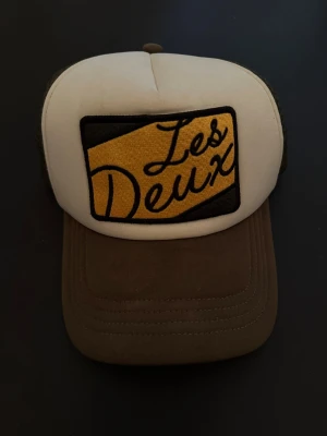 Les Deux brun och vit keps - Säljer en snygg keps från Les Deux med broderad patch framtill. Kepsen är brun och vit med mesh på sidorna och bak, samt en gul och svart detalj på framsidan. Perfekt för dig som gillar streetwear och vill ha en cool accessoar.