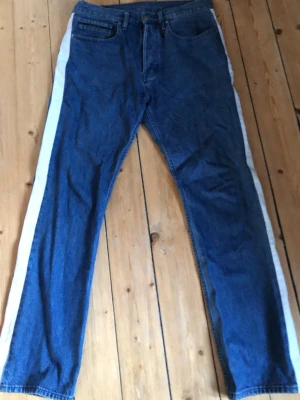 Calvin Klein jeans med vit sidoremsa - Säljer ett par Calvin Klein jeans i klassisk blå denim med raka ben och vit sidoremsa längs utsidan. Modellen har normal passform och fem fickor. Perfekt för dig som vill ha en unik touch på dina jeans. Materialet är slitstark bomullsjeans. 