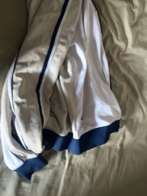 Vit och beige ziptröja från Ellesse - Säljer en snygg ziptröja från Ellesse i vitt och beige med marinblå detaljer. Tröjan har dragkedja framtill, ribbade muddar och tunna marinblå ränder längs ärmarna. Klassisk logga på bröstet och fickor med dragkedja. Perfekt till streetwear eller sportig stil.