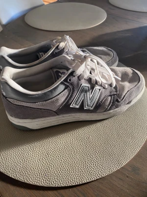 Grå New Balance sneakers 8.5 US - Säljer ett par grå New Balance sneakers med vit sula och vita snören. Skorna har en klassisk rund tå och är tillverkade i mocka och syntetmaterial. Stort N på sidan och New Balance-logga på hälen. Perfekta för dig som gillar stilrena och bekväma sneakers.
