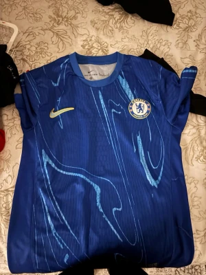 Chelsea  - Bilderna visar en blå Chelsea FC fotbollströja från Nike med gula och vita detaljer, men inga skor syns på bilderna.