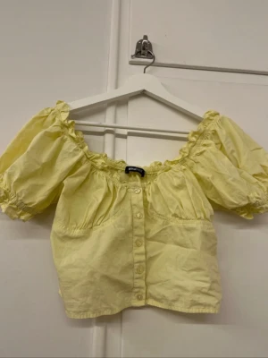 Gul topp - Säljer en ljusgul blus från Gina Tricot med puffiga korta ärmar och volangdetaljer vid halsringningen. Blusen har knappar framtill och är croppad i modellen. Perfekt för sommarens alla tillfällen! Croppad