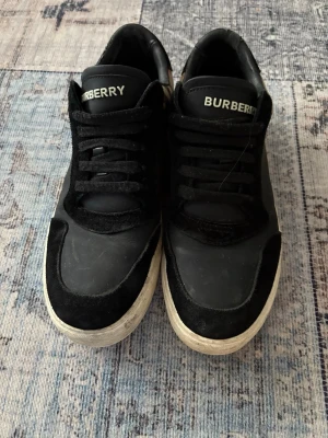 Burberry svarta sneakers med rutig detalj - Snygga svarta sneakers från Burberry med klassisk rutig panel på sidan. Skorna har snörning, platt sula och rund tå. Materialet är en mix av skinn och mocka, med vit yttersula och Burberry-logga på plösen. Perfekt för dig som gillar stilrena och lyxiga detaljer. St 42, men passar 42,5
