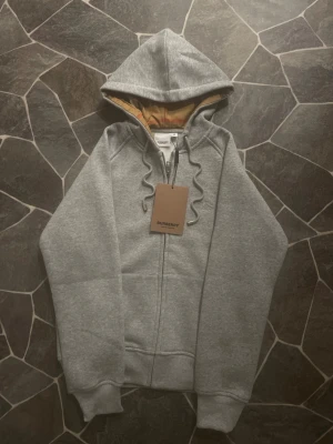 Grå hoodie från Burberry med dragkedja - Snygg grå hoodie från Burberry med klassisk logga broderad på bröstet och ikoniskt rutigt foder i huvan. Tröjan har snörning vid huvan, ribbade muddar och en rejäl dragkedja framtill. Perfekt för dig som gillar stilrena och lyxiga streetwear-plagg. Plagget sitter som S - M.