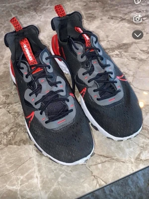 Nike React sneakers svart/röd - Nike React sneakers i svart och rött med grå detaljer och vit sula med svarta mönster. Snygg design med röd swoosh och häldetalj, samt snörning och mesh-överdel för bra ventilation. Skorna har rund tå och platt sula, perfekt för dig som gillar sportig stil.