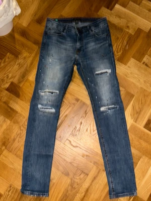 Blå jeans med slitningar och färgstänk - Säljer ett par blå jeans från Shop&Chill med coola slitningar och färgstänk på både fram- och baksida. Jeansen har fem fickor, knappgylf och klassisk passform. Materialet är jeans med snygg tvätt och detaljer som ger en edgy vibe.