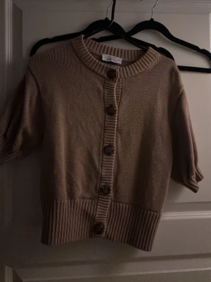 Beige stickad kofta från & Other Stories - Säljer en beige stickad kofta från & Other Stories med korta ärmar och stora knappar framtill. Koftan har ribbade muddar vid hals, ärmslut och nederkant. Perfekt att slänga över en topp för en chill look.