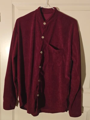 Vinröd manchester henley med guldknappar - Säljer en vinröd henley-skjorta i mjuk manchester från CA Prods Sportswear. Skjortan har lång ärm, ståkrage och snygga guldknappar med stjärndetalj. En bröstficka på framsidan och loose passform gör den chill och enkel att styla.