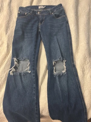 Blå jeans med stora hål på knäna - Säljer ett par blå jeans med riktigt stora slitna hål över båda knäna för en edgy look. Klassisk femficksmodell med raka ben och normal passform. Perfekta för dig som gillar streetstyle och vill ha ett par statement-jeans.
