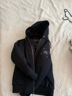 Svart pufferjacka från Moose Knuckles - Svart  jacka från Moose Knuckles med huva och quiltad insida. Jackan har dragkedja framtill, snörning vid huvan och ett snyggt logomärke på ärmen. Materialet är polyester och jackan har en loose passform som är perfekt för kalla dagar.