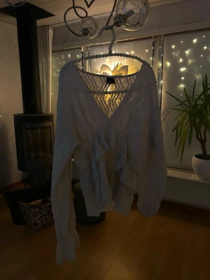 Vit blus med volang och v-ringning - Säljer en vit blus med v-ringad hals och stora volangdetaljer framtill. Blusen har långärmade ärmar och ett krinklat, lätt transparent tyg som ger en cool och unik look. 