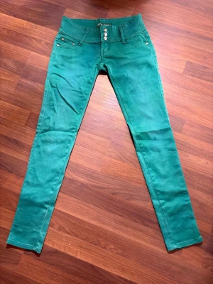 Gröna skinny jeans från R-Ping, stl 27 - Snygga gröna jeans från R-Ping i storlek 27. Modellen är skinny med låg midja och klassiska fem fickor. Materialet är 95% bomull och 5% elastan för en bekväm och stretchig passform. Framtill finns tre silverfärgade knappar och dragkedja. OBS dragkedjan är trasig annars bra skick 