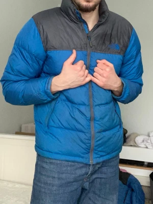 Blå dunjacka från The North Face M - Blå och mörkgrå dunjacka från The North Face i storlek M. Jackan har hög krage, lång ärm och dragkedja framtill. Fylld med dun för extra värme och har två sidofickor med dragkedja. Perfekt för kalla dagar och har en clean, sportig look.