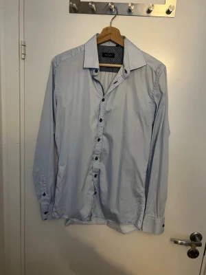 Ljusblå skjorta från Sand Copenhagen - Snygg ljusblå skjorta från Sand Copenhagen i storlek 41. Skjortan har klassisk krage, svarta knappar och lång ärm. Insidan av kragen har ett mörkblått prickigt mönster som ger en cool kontrast. Perfekt för dig som gillar stilrena detaljer. Aldrig använd, inga defekter, skriv vid frågor och funderingar.
