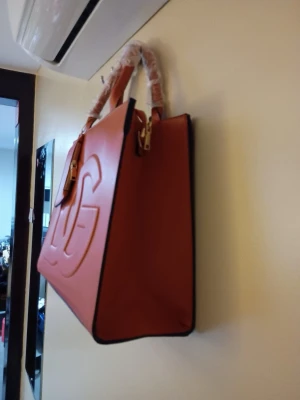 Handväska  - Snygg orange handväska från med stor präglad logga på framsidan och matchande liten plånbok med dragkedja. Väskan har två robusta handtag och är tillverkad i slätt skinnmaterial. Perfekt för dig som gillar stilrena och trendiga accessoarer.