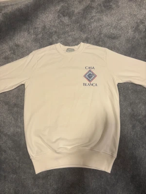 Vit sweatshirt från Casablanca S - Vit sweatshirt från Casablanca Tennis Club i storlek S. Tröjan har ett tryckt emblem med blått och rött mönster samt texten 'Casa Blanca' på bröstet. Rund hals och långa ärmar, tillverkad i mjuk bomull. Perfekt för en chill och sportig look.
