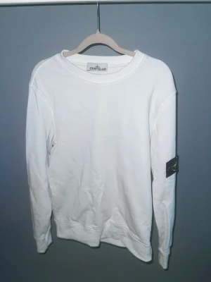 Vit sweatshirt från Stone Island - Vit långärmad sweatshirt från Stone Island med klassisk rund hals och ikonisk svart patch med gul broderad logga på vänster ärm. Tröjan är i mjuk bomull och har en clean, normal passform som funkar till många olika stilar.