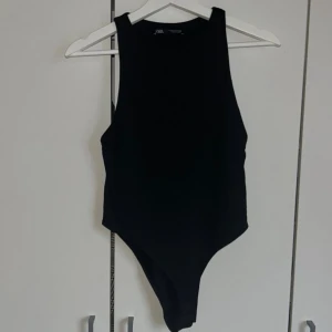 Svart ärmlös bodysuit från Zara - Säljer en svart, stilren bodysuit från Zara med hög hals och ärmlös design. Perfekt att styla med jeans eller kjol för en clean look. Materialet är mjukt och stretchigt för en bekväm passform.