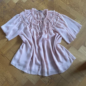 Ljusrosa volangblus med knyt - Supersöt ljusrosa blus från Zara med volangdetaljer och knyt framtill. Blusen har korta, vida ärmar och är tillverkad i ett tunt, flowy material som ger en luftig känsla. Passar större/mindre storlekar