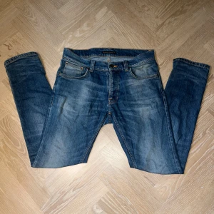 Blå jeans från Nudie Jeans, 30/30 - Säljer ett par klassiska blå jeans från Nudie Jeans i storlek 30/30. Jeansen har en snygg tvätt med slitningar och orangea sömmar. Modellen är rak med normal passform och tillverkad i mjukt jeanstyg. Perfekta för dig som gillar en tidlös look.