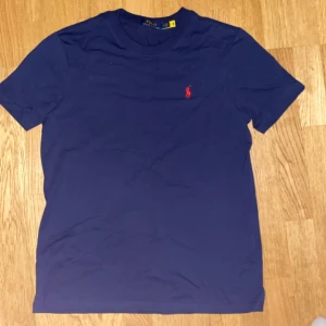 Blå t-shirt från Polo Ralph Lauren - Snygg marinblå t-shirt från Polo Ralph Lauren med klassisk röd broderad logga på bröstet. T-shirten är i mjuk bomull och har en normal passform med korta ärmar och rund hals. Perfekt för en clean och stilren look.