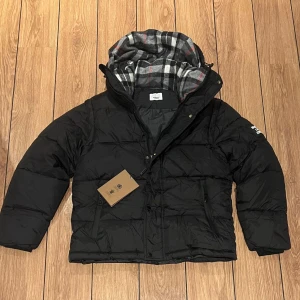 Svart pufferjacka från Burberry M - Svart pufferjacka från Burberry med avtagbara ärmar och huva fodrad i klassiskt rutigt mönster. Jackan har dragkedja, tryckknappar och två fickor framtill. Stor Burberry-patch på ena ärmen. Yttermaterial i bomull, innerfoder i bomull och viskos. Storlek M men passar perfect för storlek S 