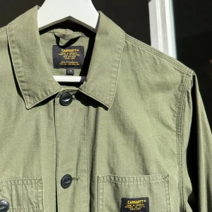 Skjortjacka från Carhartt - Säljer en grön skjortjacka från Carhartt. Jackan är i 100% bomull, har fyra stora fickor framtill och stängs med knappar. Klassisk krage och diskret Carhartt-logga på bröstfickan. Inköpt i Biarritz.