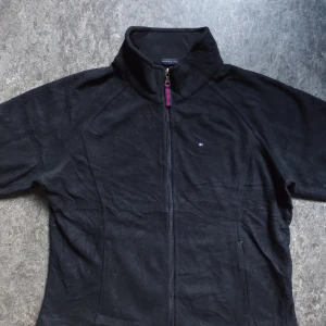 Fleecejacka från Tommy Hilfiger - Svart fleecejacka från Tommy Hilfiger med hel dragkedja och hög krage. Jackan har diskret logga på bröstet och snygga detaljer i rött och blått vid dragkedjan. Perfekt för kyliga dagar och enkel att matcha med allt.                                                                      OBS det finns små fläckar i insidan som inte syns  i utsidan men ändå något man får informera om
