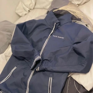 Blå vindjacka från Race Marine - Snygg marinblå vindjacka från Race Marine med vita detaljer och dragkedja framtill. Jackan har hög krage, långa ärmar och två fickor med vita dragkedjor. Perfekt för blåsiga dagar och enkel att matcha med det mesta.