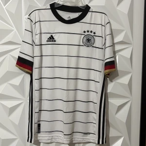 Tysklands matchtröja Adidas herr - Vit fotbollströja från Adidas med svarta horisontella ränder, svart krage och Tysklands landslagsmärke på bröstet. Ärmarna har detaljer i svart, rött och guld. Tillverkad i lätt och ventilerande material, perfekt för match eller träning.