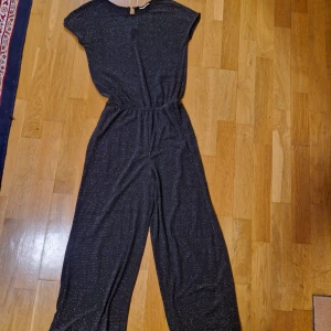 Glittrig svart byxdress från KappAhl - Snygg svart byxdress från KappAhl med glittriga detaljer. Vida ben och resår i midjan ger en avslappnad men cool look. Materialet är mjukt och stretchigt. Rund halsringning och korta ärmar med en knapp bak.