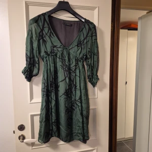 Grön mönstrad klänning från Zara Woman - Grön kortklänning från Zara Woman med svart abstrakt mönster. Klänningen har v-ringning, knyt under bysten och trekvartsärmar med resår vid ärmslut. Tillverkad i ett lätt, flowy silke som ger en avslappnad och snygg siluett.