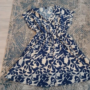 Blåvit omlottklänning med bladmönster - Snygg omlottklänning från H&M i blått och vitt med ett stort bladmönster. Klänningen har korta ärmar, v-ringad hals och knyt i midjan för en skön passform. Perfekt för dig som gillar en fräsch och somrig stil.