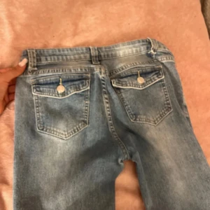 Ljusblå jeans med fickdetaljer - Säljer ett par ljusblå jeans med snygga slitningar och coola ficklock med knapp bak. Klassisk femficksmodell med dragkedja och knapp framtill. Jeansen har en rak passform och är perfekta för en avslappnad stil.