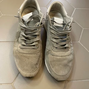 Grå mockasneakers från Philippe Model - Snygga sneakers från Philippe Model i grå mocka med vita detaljer på hälen. Skorna har klassisk snörning, rund tå och platt sula. Perfekta för dig som gillar stilrena och neutrala färger. Philippe Model-logga på sidan ger en exklusiv touch. Fråga om mer bilder om du är intresserad