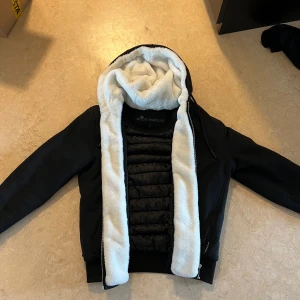 Moose Knuckles jacka - Säljer en svart classic Bunny från Moose Knuckles med vit teddyfoder i huvan och insidan. Jackan har dragkedja, ribbade muddar och ikonisk logga på ärmen. Materialmix av polyester och syntet, perfekt för kalla dagar. Snygg och modern look med quiltad insida. Äkta kvitto finns köpt förra året på johnells. Pris kan diskuteras vid snabb affär
