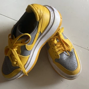 Nike Jordan 1 Low gul och grå - Nike Jordan 1 Low sneakers i en fet gul och grå färgkombo med vit sula och platt profil. Skorna har snörning, rund tå och klassisk Jordan-logga på hälen. Ovandelen är i skinn och detaljerna är riktigt snygga med kontrasterande färger och tydliga sömmar.