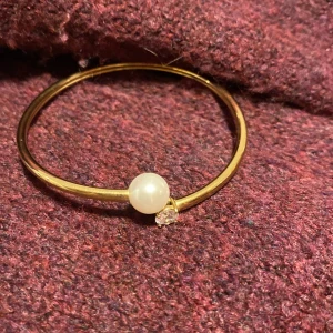 Guldfärgat armband med pärla från Edblad - Stilrent armband från Edblad i guldfärgad metall med en vit pärla och en gnistrande sten på öppningen. Enkel och modern design som ger en lyxig känsla till din outfit. Perfekt accessoar för dig som gillar eleganta detaljer.