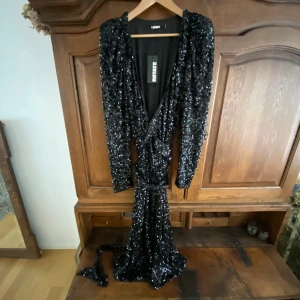 Svart paljettklänning från Rotate - Maxad svart långklänning från Rotate, täckt av glittriga paljetter. Djup v-ringning framtill, markerade axlar och lång ärm. Klänningen har ett bälte i midjan och en snygg slits för extra edge. Perfekt för dig som vill sticka ut med en glamorös look.