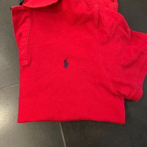 Röd pikétröja från Polo Ralph Lauren - Klassisk röd pikétröja från Polo Ralph Lauren med blå broderad logga på bröstet. Tröjan har korta ärmar, krage och knappar framtill. Tillverkad i bomull och har en snygg, enkel design som passar perfekt till jeans eller shorts.