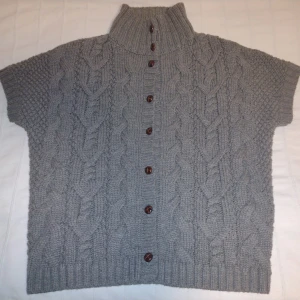 Grå/beige stickad väst/kofta med knappar och krage  - Mysig grå/beige/brun stickad kofta från Hampton Republic med korta ärmar och hög ribbad krage. Koftan har ett snyggt flätmönster och bruna knappar framtill. Perfekt för lager-på-lager och chill höst- och vinterstil. Storlek 36/38. Är lite nopprig.