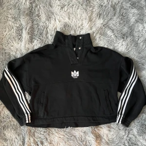 Svart Adidas tröja med halv zip - Svart Adidas tröja med klassiska vita ränder längs ärmarna och broderad logga framtill. Hög krage med tryckknappar och halv zip. Långärmad och i mjuk bomull, perfekt för en sportig och chill look.